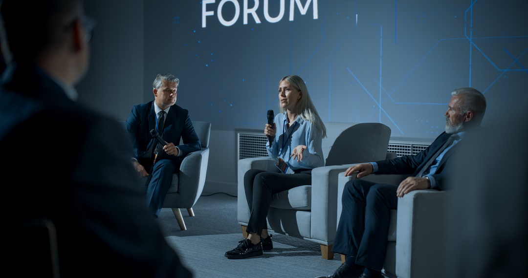 Conference speaker panel with female speaker talking into a microphone with men on either side