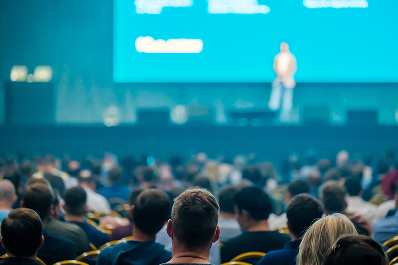 Audience seated in a conference hall facing a presenter on a large stage